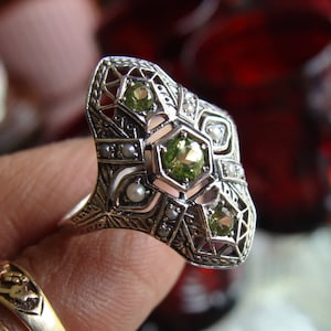 May include: A silver-toned ring featuring an elongated, openwork filigree design. The centerpiece is adorned with three round, faceted olive-green stones and several small, round white pearls set within the intricate metal lattice structure.
