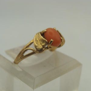 Op de afbeelding: Een gouden ring met een ovale, koraalkleurige steen. De band van de ring heeft aan weerszijden van de steen een bladerontwerp. De ring wordt gepresenteerd op een heldere, rechthoekige standaard tegen een witte achtergrond. De ring is een sieraad.