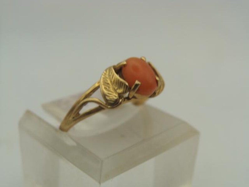 Op de afbeelding: Een gouden ring met een ovale, koraalkleurige steen. De band van de ring heeft aan weerszijden van de steen een bladerontwerp. De ring wordt gepresenteerd op een heldere, rechthoekige standaard tegen een witte achtergrond. De ring is een sieraad.