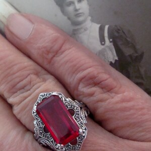 May include: A silver ring with a large, rectangular, deep red gemstone. The ring has an ornate, filigree design around the stone. A vintage black and white photograph is in the background.