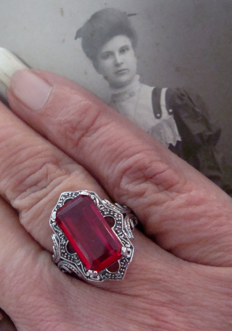 May include: A silver ring with a large, rectangular, deep red gemstone. The ring has an ornate, filigree design around the stone. A vintage black and white photograph is in the background.