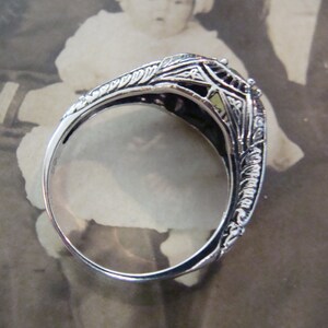 May include: A silver filigree ring with intricate floral and geometric designs. The ring's band is thin and polished, and the setting features a small, light-colored stone. The ring is set against a vintage photograph background.