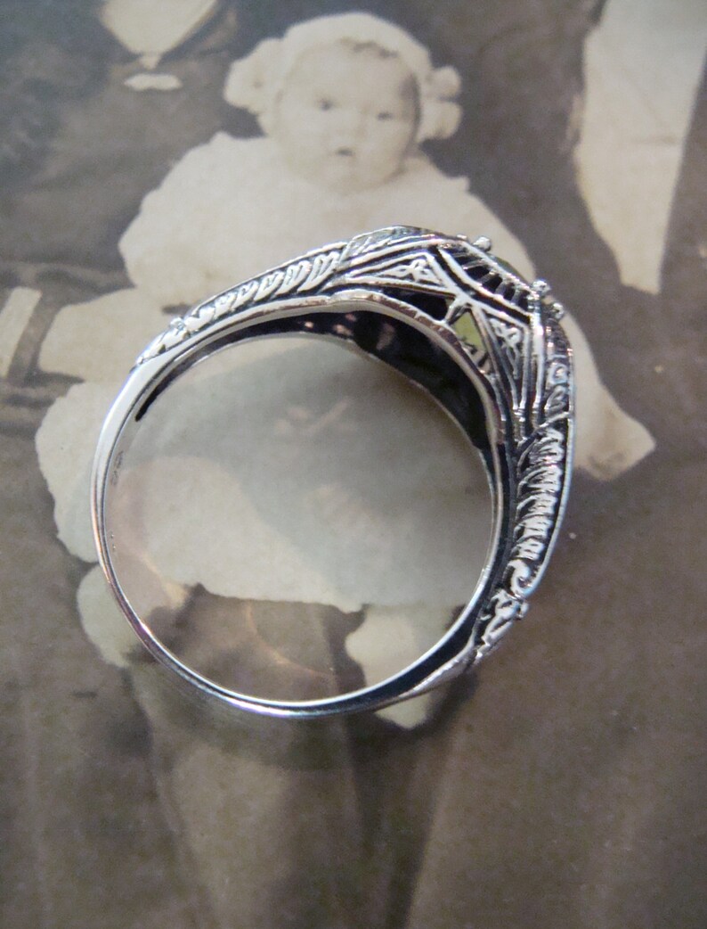 May include: A silver filigree ring with intricate floral and geometric designs. The ring's band is thin and polished, and the setting features a small, light-colored stone. The ring is set against a vintage photograph background.