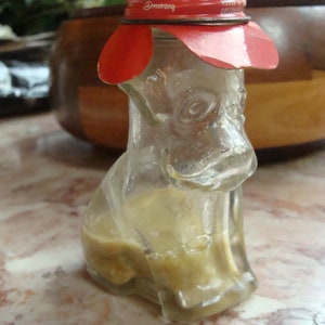 May include: A clear glass dog-shaped bottle with a red and white cap. The bottle is partially filled with a light brown liquid. The cap has the text "@mstrong" printed on it. The bottle is sitting on a marble surface.