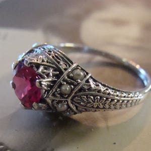 May include: A silver-toned ring featuring a round, faceted, deep pink center stone held in a decorative filigree setting. The band is adorned with intricate metal leaf patterns and small, round, white pearls embedded along the sides.
