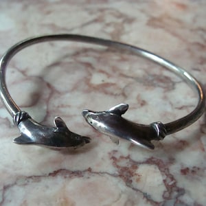 May include: A silver-toned bracelet featuring two dolphin figures at the ends. The dolphins are detailed with fins and facial features, and the bracelet has a simple, curved design. The jewelry is displayed on a marble surface.