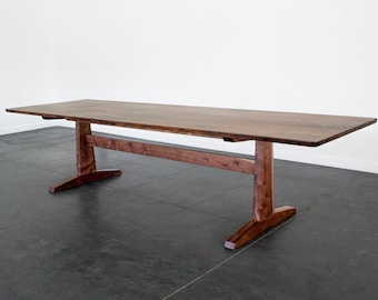 Könnte beinhalten: Ein großer, rechteckiger Esstisch aus Holz mit dunkelbraunem Finish. Der Tisch hat ein Gestell mit zwei vertikalen Stützen, die durch einen horizontalen Balken verbunden sind. Der Tisch ist ca. 244 cm lang.