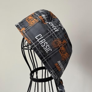 May include: A gray and black plaid bandana with orange motorcycle graphics and the words "CLASSIC" and "MOTORCYCLE MANIA". The bandana is displayed on a black metal stand.