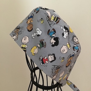 May include: A gray fabric scrub hat with a tie, featuring a repeating pattern of Peanuts characters in various poses. The characters are in black, white, yellow, blue, and red.