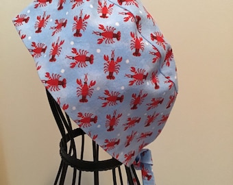 May include: A light blue scrub hat with a white polka dot pattern and red lobster print. The hat has a tie that can be tied at the back of the neck.
