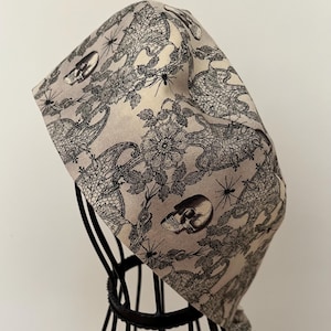 May include: A beige and black skull and spider patterned head covering. The fabric features a repeating pattern of skulls, spiders, and floral lace designs. The head covering is displayed on a black wire mannequin head.