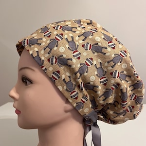 May include: A beige surgical scrub cap featuring a repeating pattern of sock monkey figures in gray, red, and white. The cap also has white button accents and gray ties for adjustment. The cap is designed to cover the head.