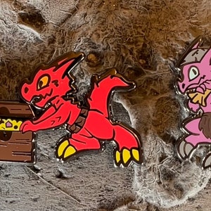 May include: Set of seven enamel pins featuring cartoon dragons in various poses. The dragons are blue, orange, red, pink, and brown. The pins are all approximately 2.5 cm in size.