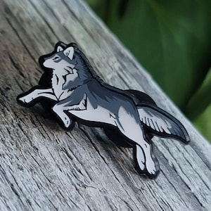 May include: A black and white enamel pin of a wolf jumping in the air. The wolf is depicted in a realistic style with a detailed fur pattern.