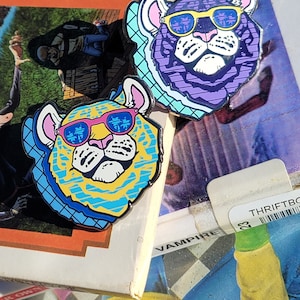May include: Two enamel pins featuring cartoon tigers wearing sunglasses. The tiger on the left is yellow and blue, while the tiger on the right is purple and blue. Both tigers have pink ears and white faces.