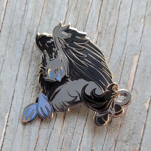 May include: A black and blue enamel pin of a mythical creature with wings, a long tail, and bright yellow eyes. The creature is depicted in a playful pose with its wings spread out.