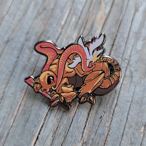 May include: A gold and orange enamel pin of a cartoon creature with a red tentacle, wings, and a stick in its hand.