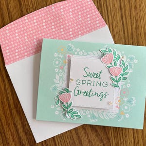 Op de afbeelding: Een lentegroetkaart met een mintgroene achtergrond en een witte envelop. De kaart heeft een bloemenontwerp met roze bloemen en de woorden "Sweet Spring Greetings". De envelop heeft een roze en wit patroon aan de binnenkant.