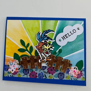 Op de afbeelding: Een handgemaakte kaart met een kleurrijke aquarel achtergrond met een zonnestralen ontwerp. De kaart heeft een cartoonvogel met een hek en bloemen. De kaart heeft het woord "HELLO" op een witte wolk geschreven.