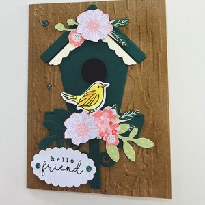 Può includere: Un biglietto d'auguri fatto a mano con una casetta per uccelli verde, un uccello giallo e fiori rosa e bianchi. Il biglietto ha un ovale bianco con il testo "hello friend" su di esso.