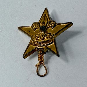 May include: A vintage gold-colored star-shaped pin with the words "BE PREPARED" inscribed on a banner. The pin features a detailed emblem and is attached to a looped wire.