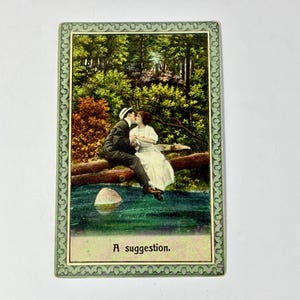 May include: Vintage postcard featuring a couple kissing on a log over a stream, surrounded by lush greenery. The card has a decorative green border and the text "A suggestion." at the bottom.