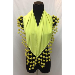 May include: A bright lime green scarf featuring a triangular shape with a deep V-neckline. The edges of the scarf are adorned with a decorative net of thin strings, each embellished with small, round, yellow fabric rosettes in a grid pattern.