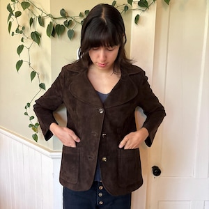 May include: A dark brown suede jacket featuring a wide collar, three front buttons, and two large patch pockets. The jacket has long sleeves and a structured fit, providing a classic look with a soft, textured finish across the entire garment.