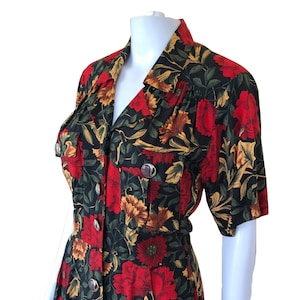 Op de afbeelding: Een zwarte, rode en gele bloemenprintjurk met een kraag, korte mouwen en knoopsluiting. De jurk heeft twee klepzakken op de borst en een getailleerde taille.