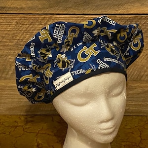 May include: Blue and gold surgical scrub hat with a repeating pattern of the Georgia Tech logo and a yellow bumblebee. The hat has a black elastic band and a white label that reads "Lil' Prints Designs".