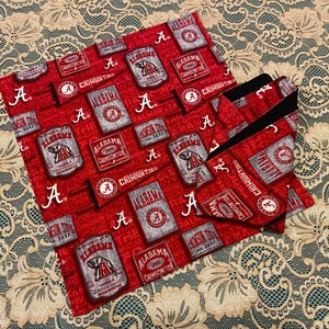 May include: Two red and black fabric squares with a pattern of the Alabama Crimson Tide logo and text. The fabric squares are on a white lace tablecloth.