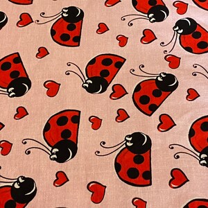 May include: Pink fabric with a repeating pattern of red ladybugs with black spots and white faces. Each ladybug is surrounded by small pink hearts.