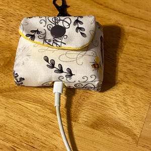 May include: A small white fabric pouch with a black button closure and a black clip. The pouch has a floral pattern with bees and the words "Be Kind" written in cursive. A white charging cable is plugged into the pouch.