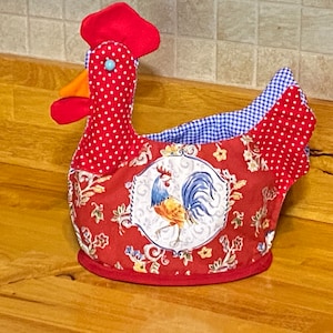 May include: A whimsical rooster-shaped kitchen item. The red fabric features white polka dots and a floral pattern. The rooster has a red comb, orange beak, and a blue eye. The interior is lined with blue and white gingham fabric.