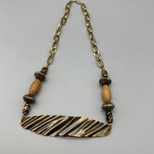 May include: A gold-toned necklace with a leaf-shaped pendant. The necklace features a chain, wooden beads, and decorative metal beads. The pendant has a textured, ridged design. The necklace is a statement piece.