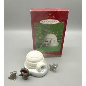 May include: A Hallmark Keepsake Ornament set featuring a white igloo, a small Inuit figure, a seal, and a husky. The box reads "Frosty Friends" and shows a similar scene. The igloo is approximately 3 inches wide.
