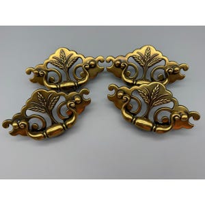 May include: A set of four brass-toned metal drawer pulls featuring an ornate, symmetrical design with a central leaf motif, curved scrollwork, and a swinging handle. Each piece has a polished finish with darker recessed areas for contrast.