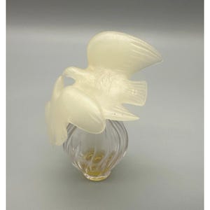 May include: A clear glass perfume bottle with a swirling design, topped with a frosted, off-white sculpture of two birds in flight. The bottle has a yellow liquid inside.