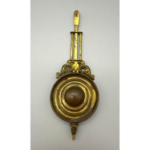 May include: Antique brass clock pendulum with a circular, ridged face and ornate detailing. The pendulum has a decorative top and a pointed bottom, showcasing a vintage aesthetic. The brass finish gives it a warm, aged appearance.