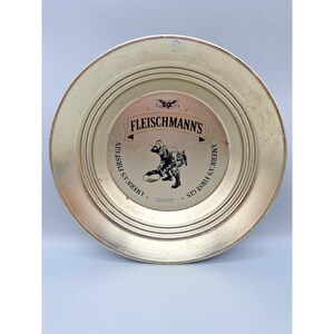 May include: A vintage, round, metal tray with a gold finish. The center features the text "FLEISCHMANN'S" above an illustration of a football player. The words "AMERICA'S FIRST GIN" are printed around the center.