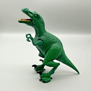 May include: A green plastic dinosaur toy with an open mouth, revealing red and yellow teeth. The toy has textured skin, black claws, and is posed in a dynamic stance. The dinosaur is a Tyrannosaurus Rex.
