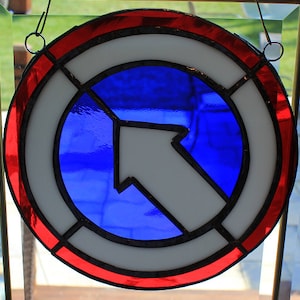 May include: Circular stained glass art piece with a red, white, and blue design. The outer ring is red, followed by a white ring, and a blue center with a white arrow pointing up and to the left. The piece is hanging.