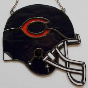 May include: Stained glass Chicago Bears football helmet with a navy blue base, a red "C" logo, and white and black detailing. The helmet is hanging from a silver chain. A decorative piece for sports fans.