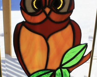 May include: Stained glass owl with a brown body, orange chest, and yellow eyes. The owl is perched on a brown branch with green leaves. The artwork is framed with a gold-colored metal.