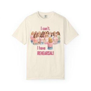 Op de afbeelding: Crèmekleurig T-shirt met de tekst "I can't. I have REHEARSAL!" in roze. Het shirt heeft een illustratie van een groep vrouwen in roze tops en een blauwe jeans. Het shirt heeft een ronde hals en korte mouwen.