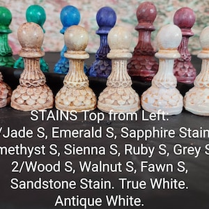 May include: A row of 12 chess pieces painted in various colors, including jade, emerald, sapphire, amethyst, sienna, ruby, grey, wood, walnut, fawn, sandstone, true white, and antique white. The chess pieces are arranged from left to right in order of their color. The text "STAINS Top from Left: 1/Jade S, Emerald S, Sapphire Stain Amethyst S, Sienna S, Ruby S, Grey S 2/Wood S, Walnut S, Fawn S, Sandstone Stain. True White. Antique White." is written below the chess pieces.