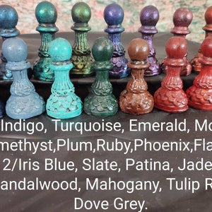 May include: A collection of 18 miniature chess pieces painted in various shades of blue, green, red, purple, and brown. The pieces are arranged in three rows on a black surface. The text below the pieces reads: "1/Indigo, Turquoise, Emerald, Moss. Amethyst, Plum, Ruby, Phoenix, Flame 2/Iris Blue, Slate, Patina, Jade, Sandalwood, Mahogany, Tulip Red Dove Grey."