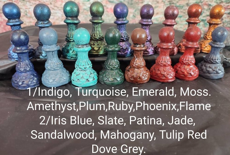 May include: A collection of 18 miniature chess pieces painted in various shades of blue, green, red, purple, and brown. The pieces are arranged in three rows on a black surface. The text below the pieces reads: "1/Indigo, Turquoise, Emerald, Moss. Amethyst, Plum, Ruby, Phoenix, Flame 2/Iris Blue, Slate, Patina, Jade, Sandalwood, Mahogany, Tulip Red Dove Grey."