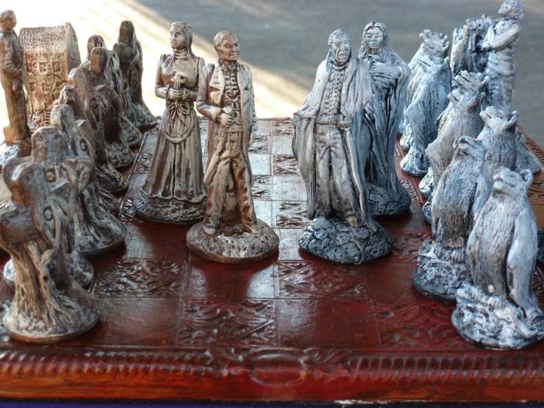 May include: A detailed chess set with intricately designed pieces. The pieces are primarily silver and bronze, depicting figures in historical attire. The chessboard is a dark, reddish-brown color with a detailed border.