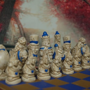 May include: A set of white ceramic chess pieces with blue accents, depicting characters from Alice in Wonderland. The pieces are arranged on a blue and brown checkered board.